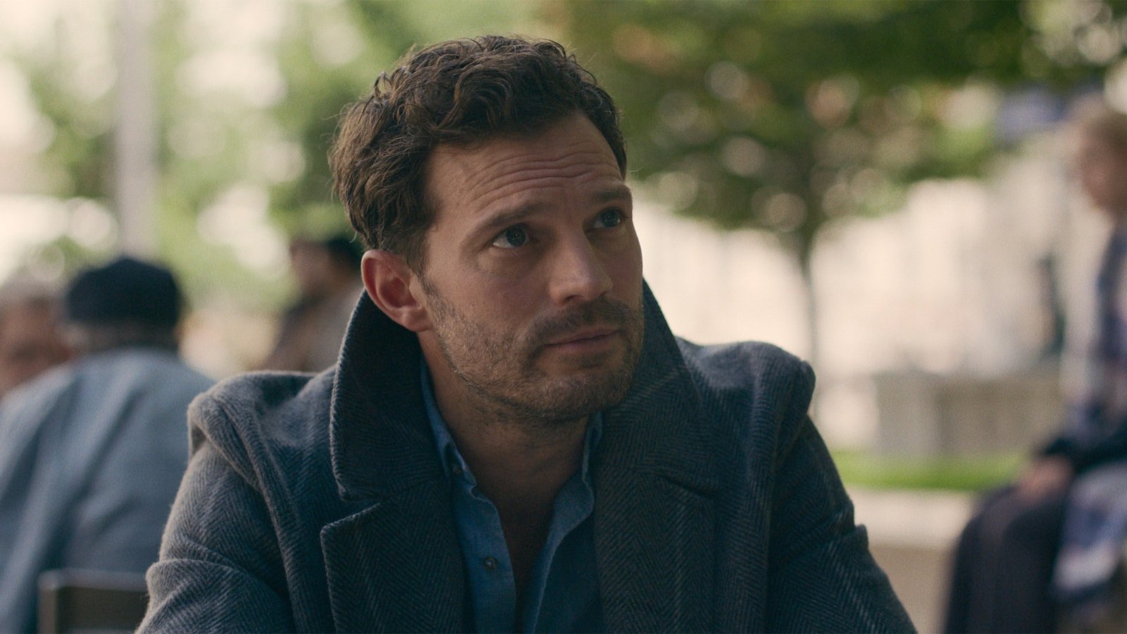 12 12 12 resmi karesinde açık havada lacivert paltosuyla bir masada oturup ciddi bir ifadeyle yana bakan Jamie Dornan