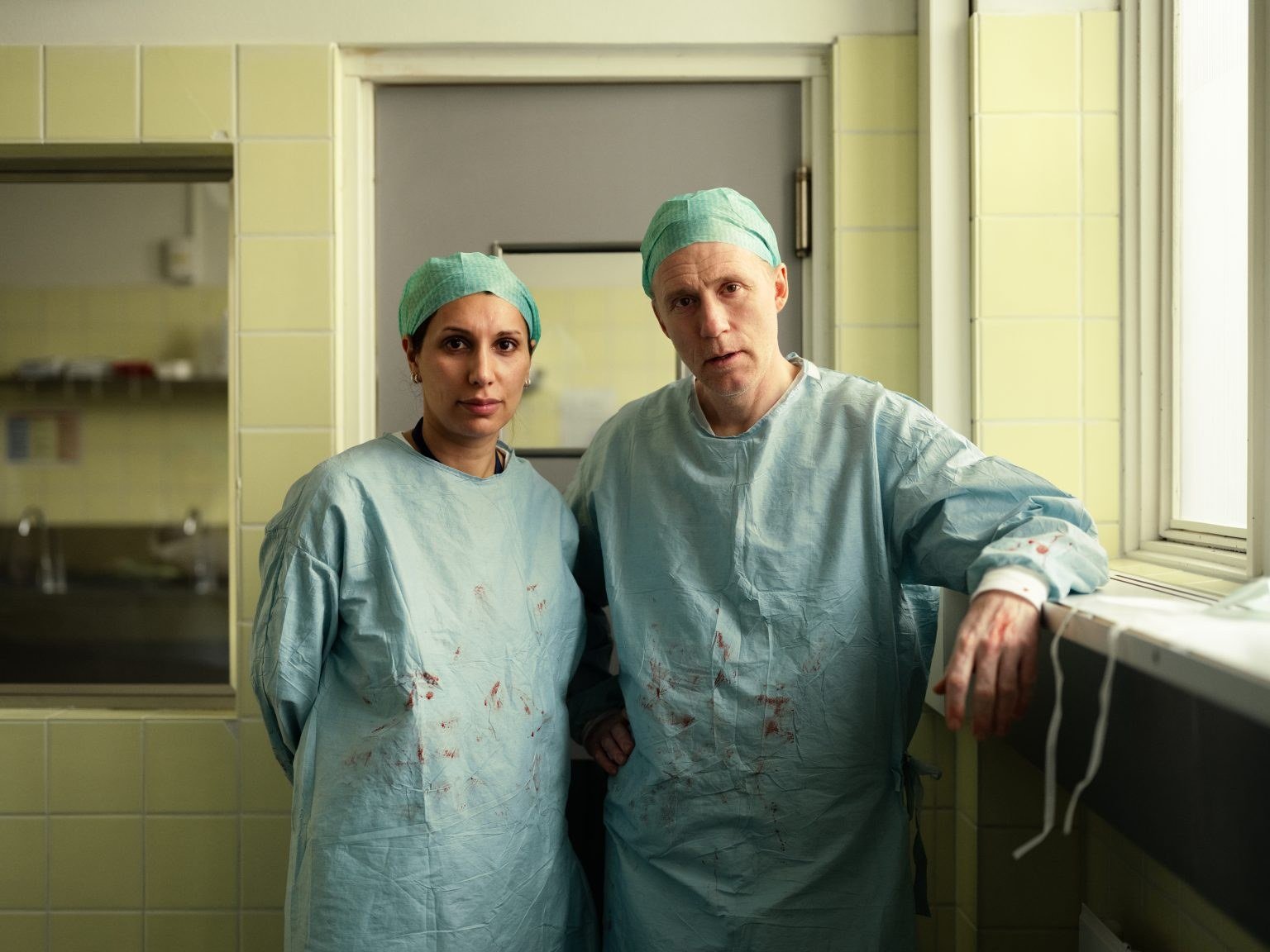Always on Call tanıtım fotoğrafında açık yeşil fayanslı bir odada cerrahi bone ve önlükle duran kadın ve erkek cerrah