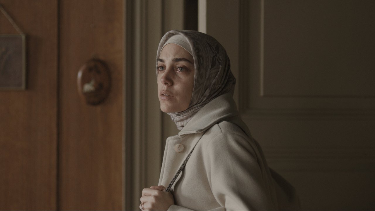 Bir Başkadır’dan Netflix karesinde başörtüsü ve açık renk paltosuyla ahşap bir kapının önünde duran Meryem rolündeki Öykü Karayel