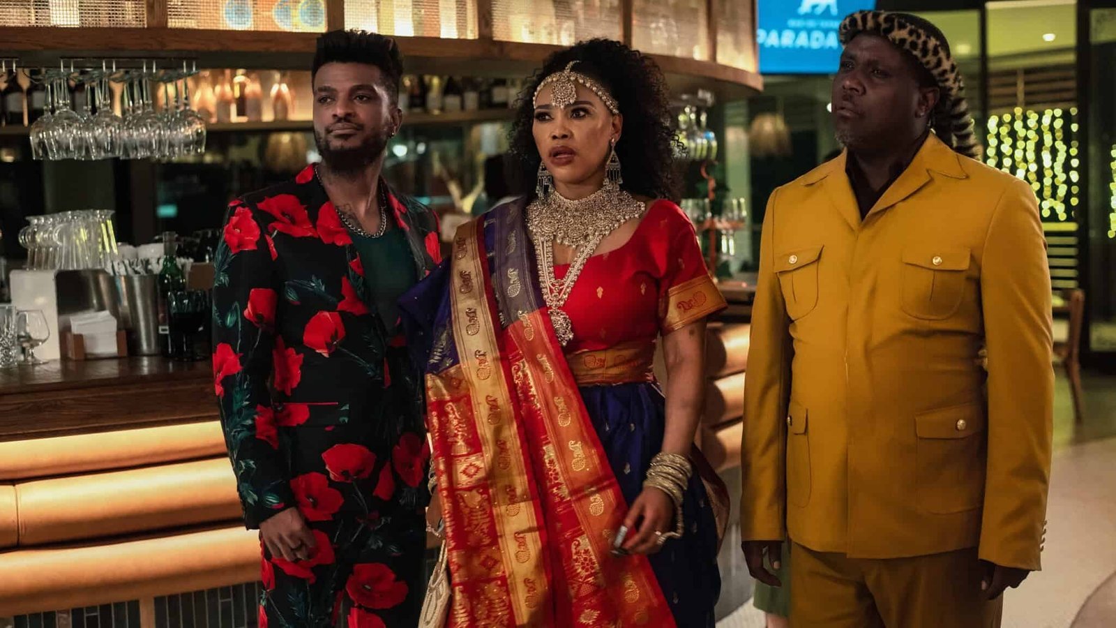 How to Ruin Diwali Netflix resmi karesinden bir restoranda renkli kıyafetleriyle duran Eazi E rolündeki Luthando Bu Mthembu, Penelope rolündeki Sonia Mbele ve Mandla rolündeki Meshack Mavuso