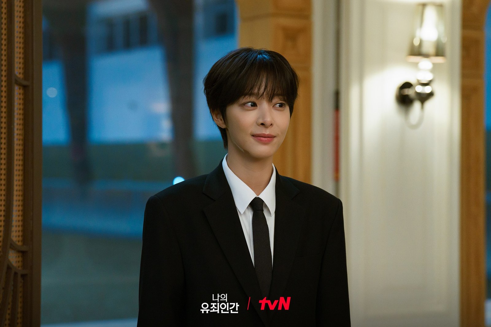 Love in Disguise tvN resmi karesinden siyah takım elbise ve kravatla bir salonda gülümseyerek duran, erkek kılığındaki Kang Jae-hee rolündeki Seol In-ah