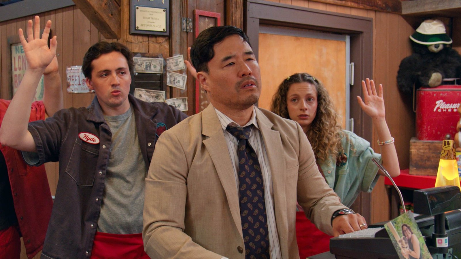 Minimum Wage Netflix resmi karesinden takım elbiseli pizzacı zinciri sahibi rolündeki Randall Park ve arkasında el kaldıran üniformalı genç çalışanlar