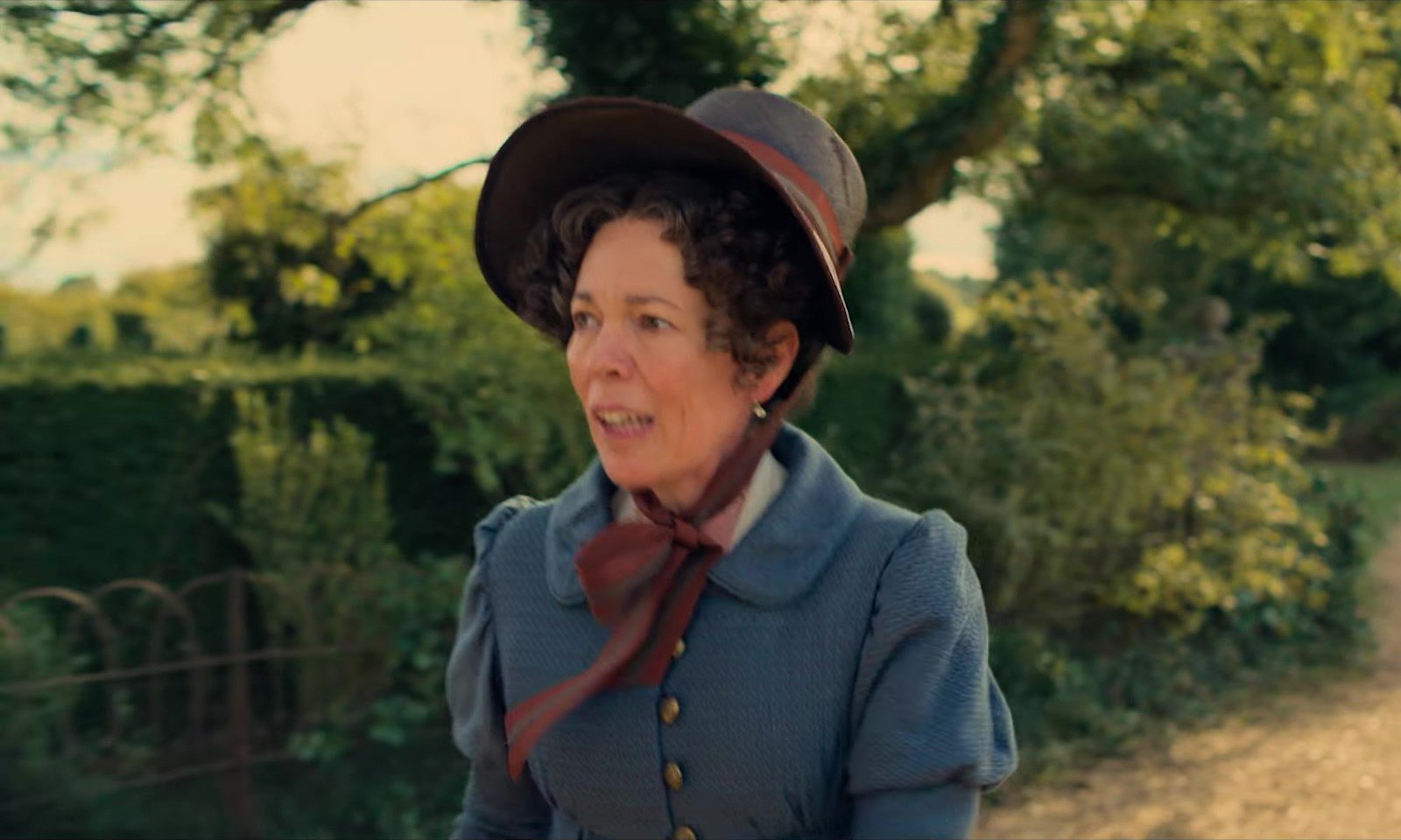 Pride and Prejudice fragmanından bahçede şapkası ve mavi ceketiyle gülümseyen Mrs. Bennet rolündeki Olivia Colman