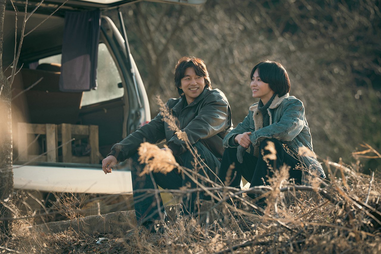 Tantara dizisinden Netflix karesinde kuru otların arasında açık bagajlı bir aracın yanında yan yana oturan Gong Yoo ve Song Hye-kyo