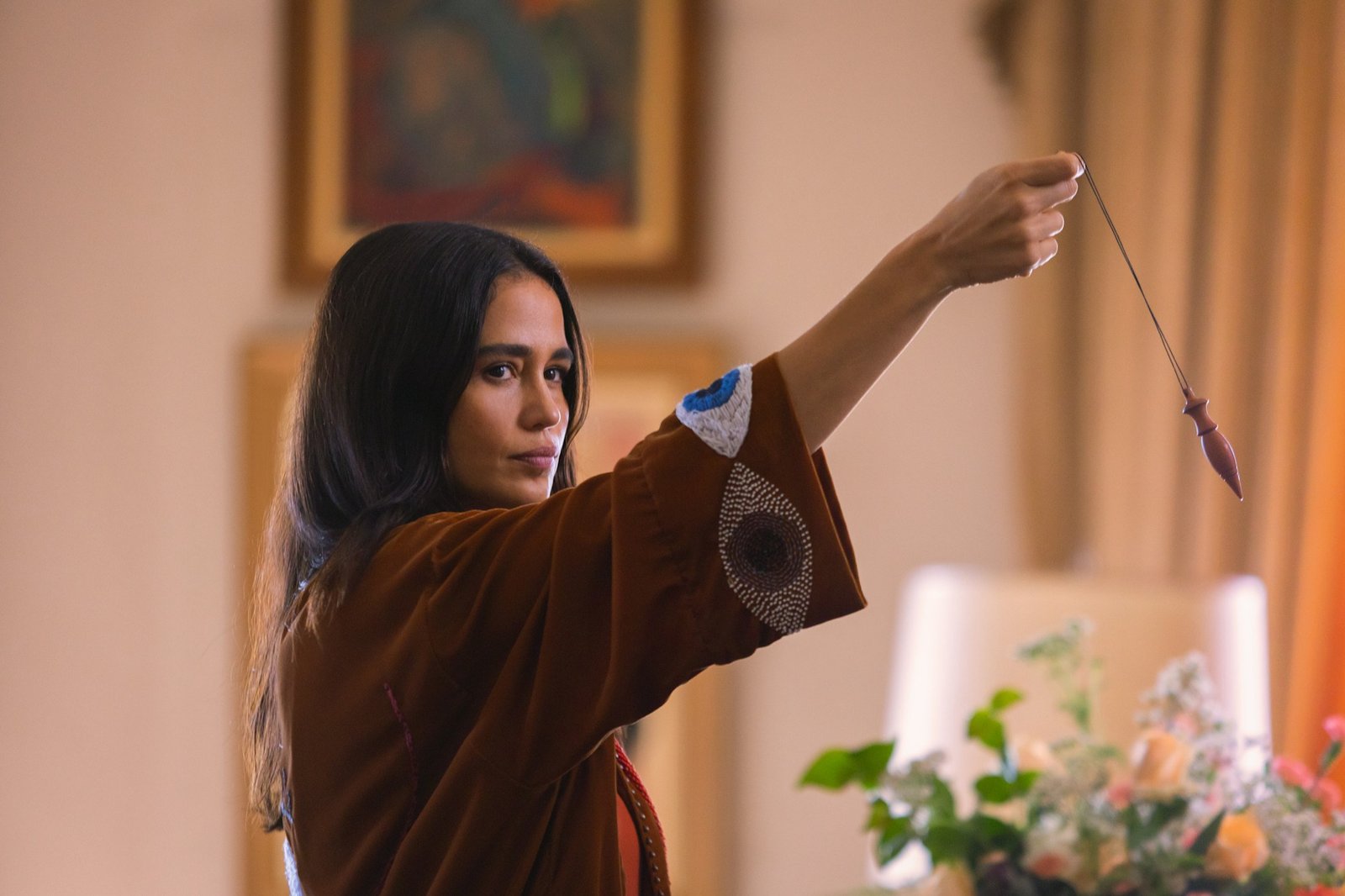 The Gift of Deceit Netflix resmi karesinden çiçekli bir salonda elindeki sarkacı havaya kaldıran spiritüel terapist Irina rolündeki Nanda Costta