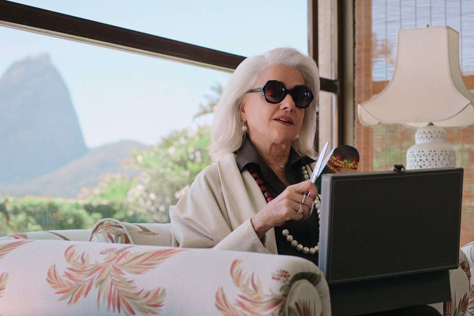 The Gift of Deceit Netflix resmi karesinden Rio de Janeiro’da Şeker Somunu Dağı manzaralı bir evde güneş gözlüğüyle oturan Lúcia Rossetti rolündeki Marieta Severo