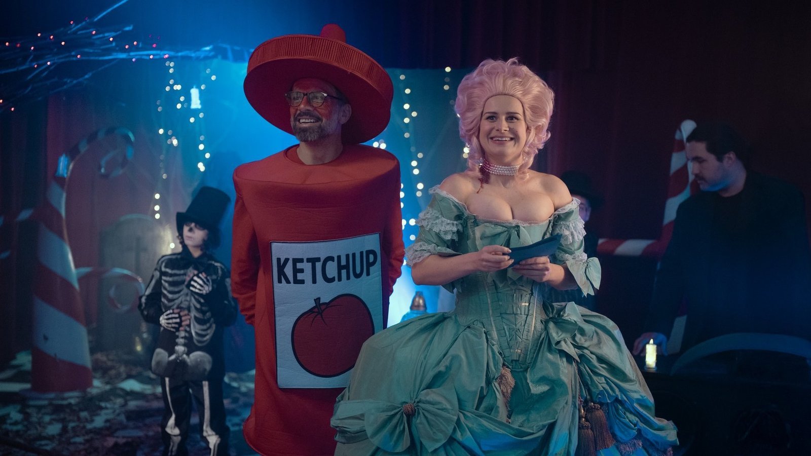 The Mothers of Penguins 2. sezon 21 Ekim’de Netflix’te: Ebeveynler bu kez kendi okullarını kuruyor 2026 – 2 The Mothers of Penguins 2. sezon resmi karesinden renkli ışıklar altında ketçap şişesi kostümlü bir kişi ve kabarık mavi elbiseli peruklu bir kadının yer aldığı kostüm partisi